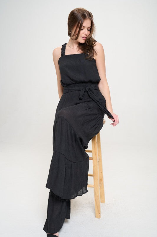 Model sitting on stool in black Chara Jumpsuit, showing flowy tiers and tie belt detail.