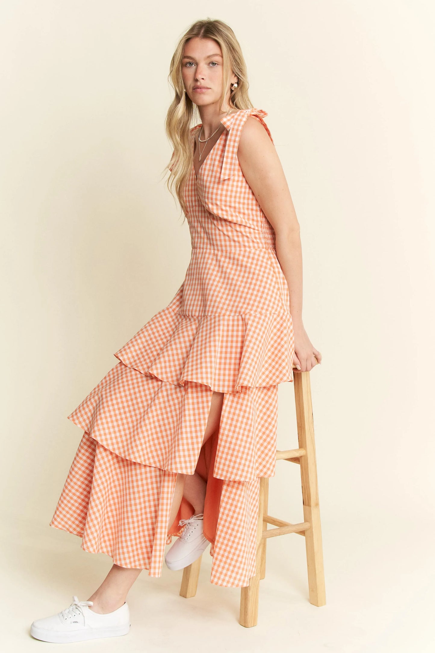 Side view of model seated in Alice Gingham Tiered Midi Dress by Jade by Jane, posing on chair with halter tie straps and flowy ruffle skirt, styled for a fresh summer look.