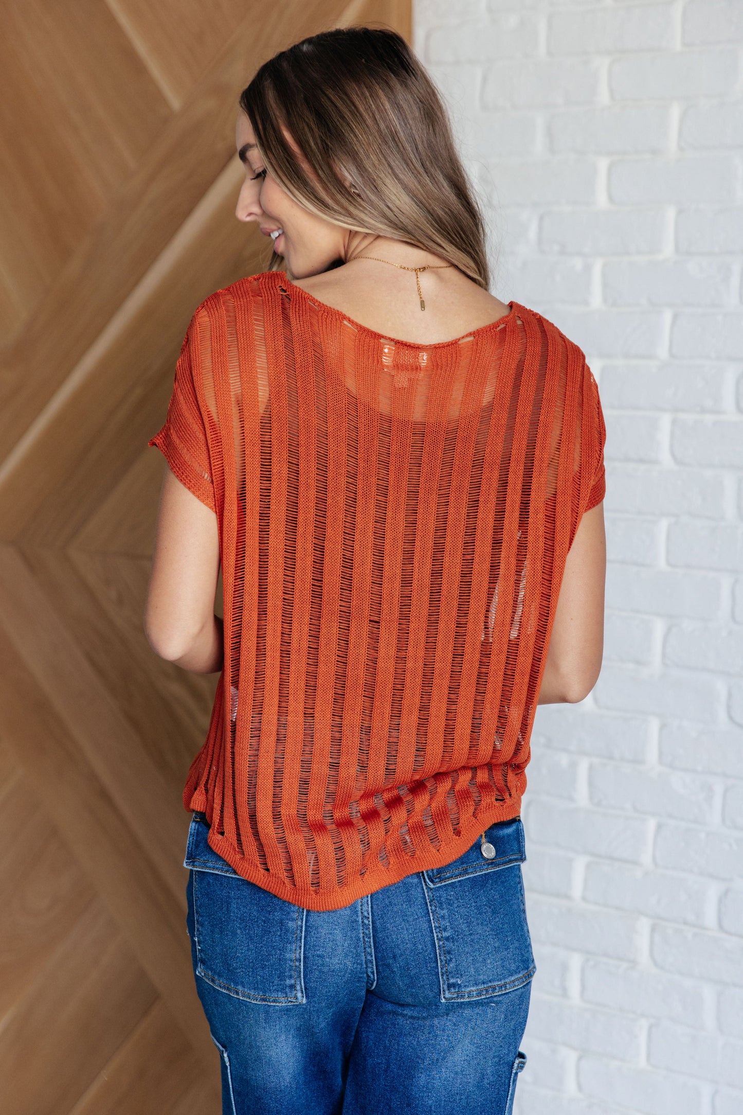 Back view of the Aaliyah Loose Knit Top in burnt orange with a wide v-neckline, short sleeves, and a lightweight open knit stripe pattern. A cozy and stylish sweater knit design.