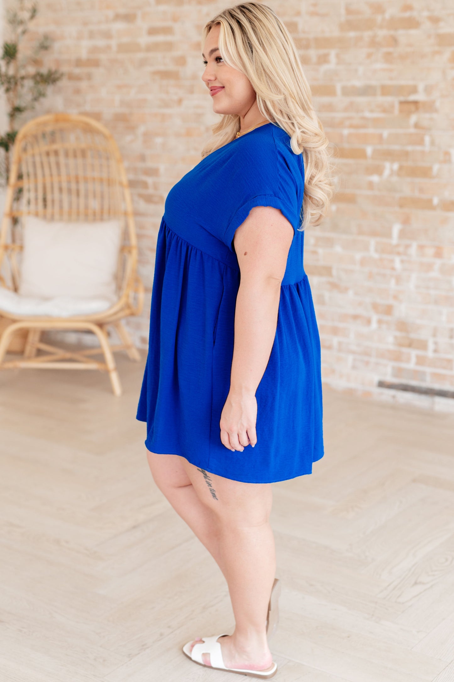 Plus size model side view of Isa V-Neck Mini Dress in blue with dolman sleeves and tacked cuffs.