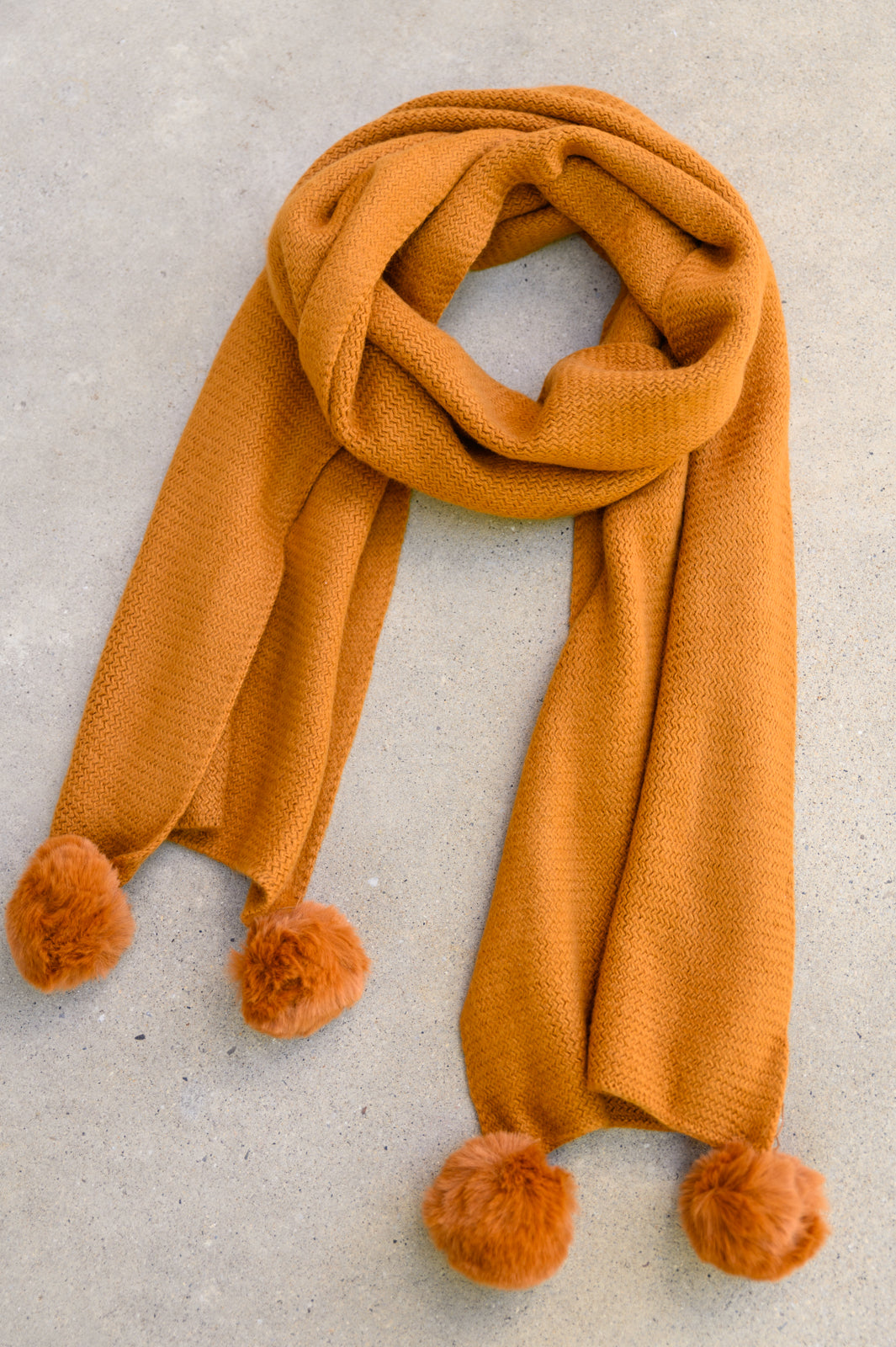 Flat lay of Ginger knitted pom pom scarf showing soft texture, warm hue, and fuzzy pom poms on each end.