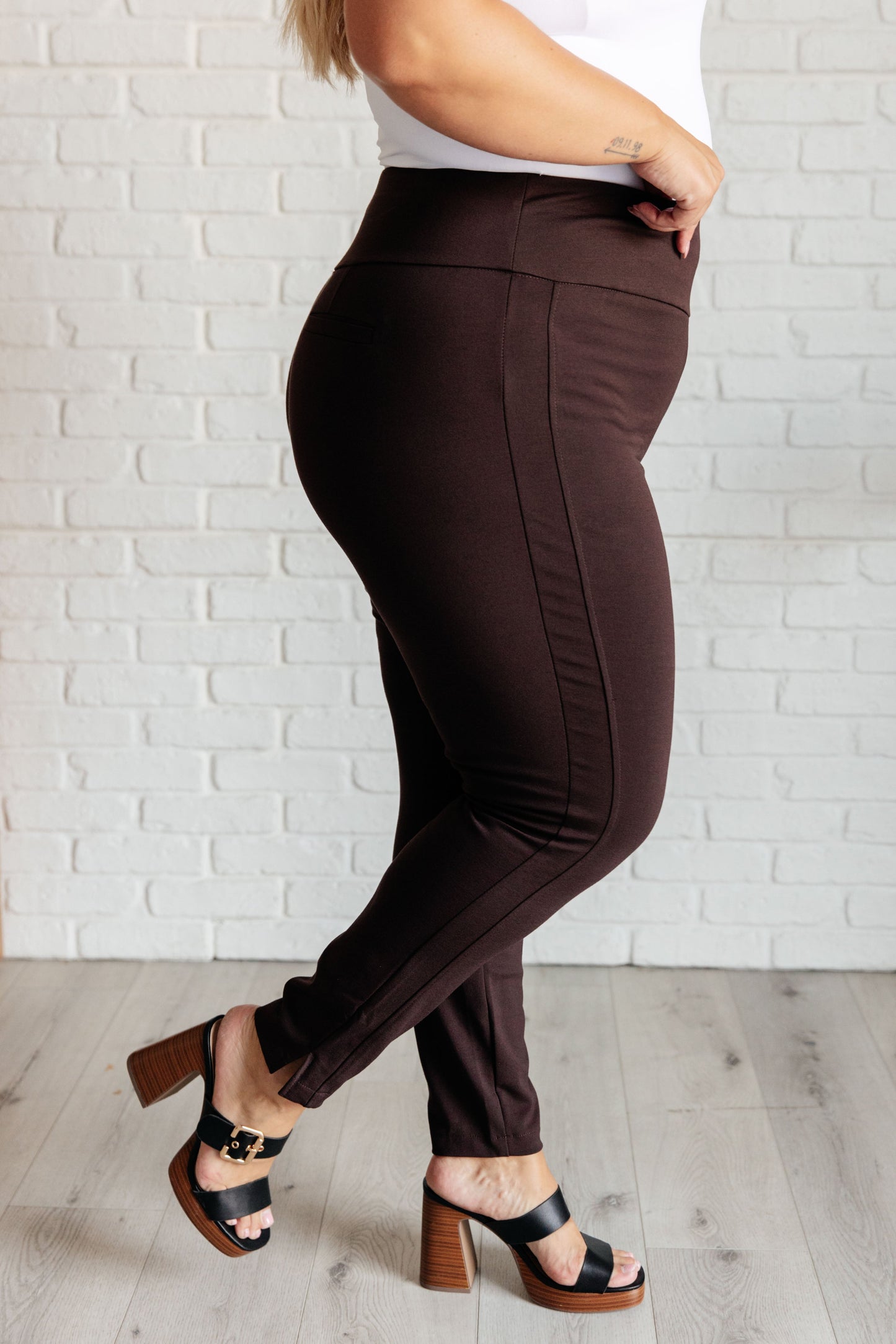 Side angle of plus-size model wearing chocolate skinny pants with tuxedo stripes and sleek silhouette.