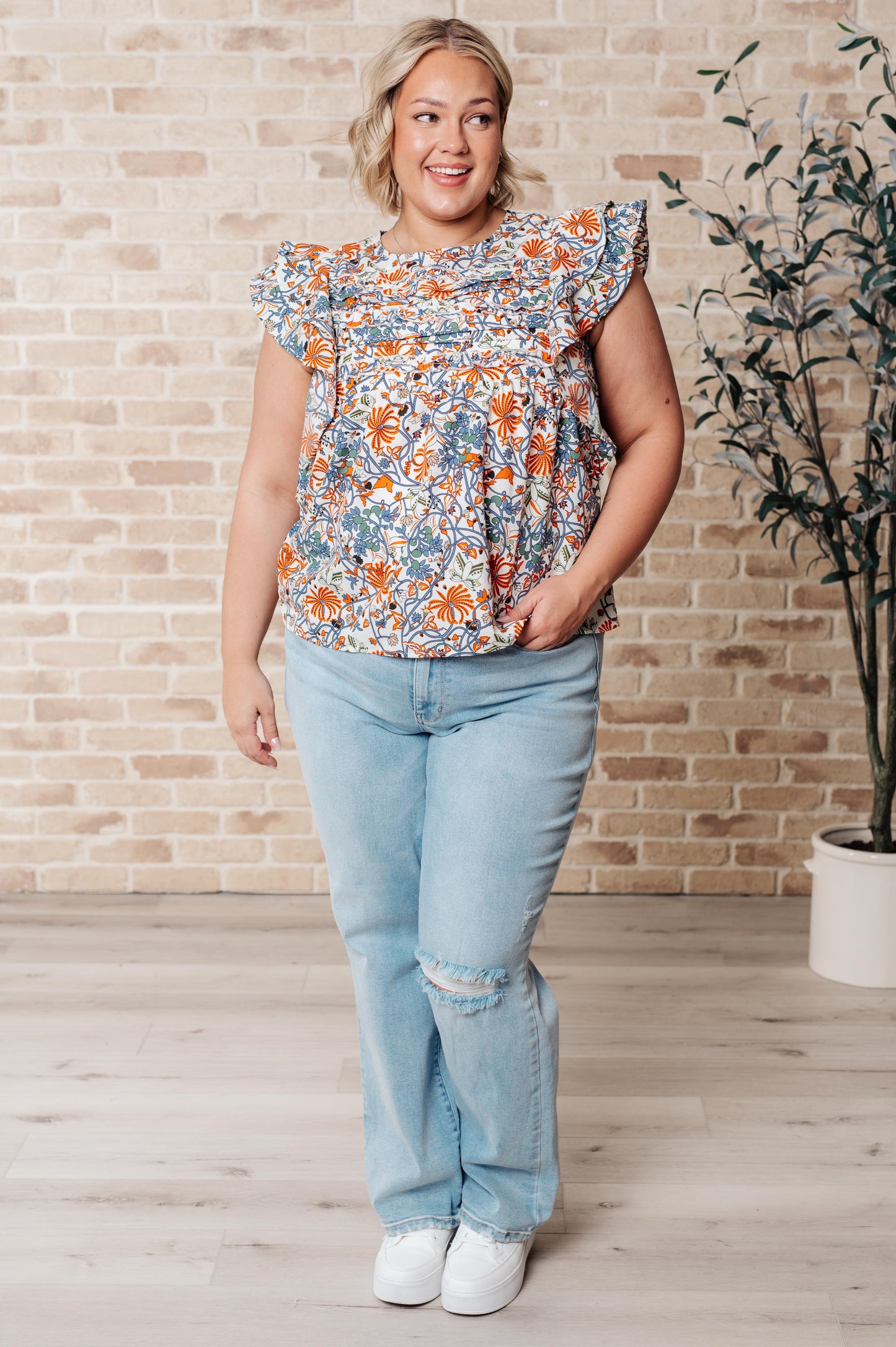Plus size model wearing Zoe Flutter Sleeve Blouse in orange and blue floral print with jeans and sneakers full body view.
