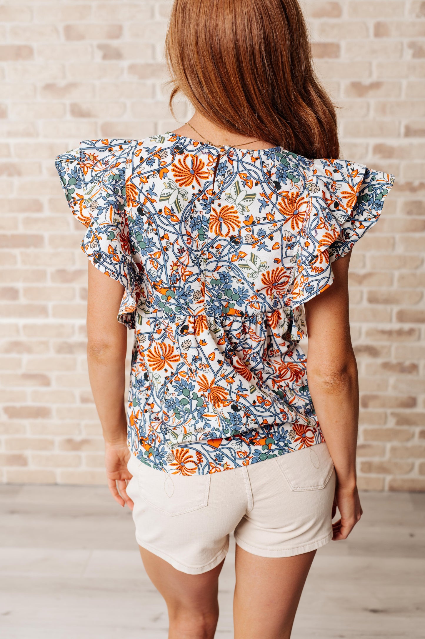Back view of Zoe Flutter Sleeve Blouse in orange and blue floral print with keyhole closure and layered flutter sleeves.