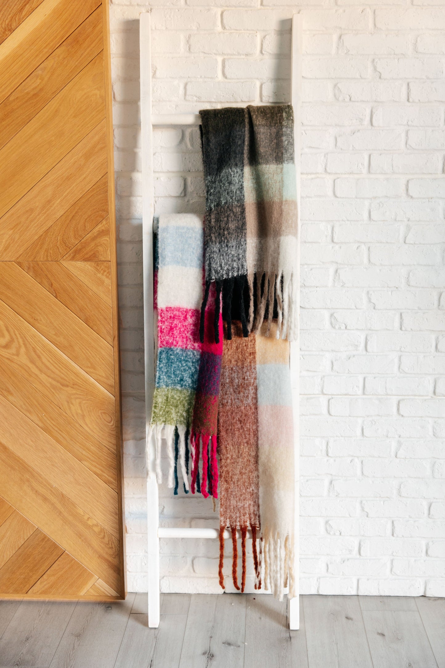 Colorful Wanderlust plaid scarves hanging on a white rack against a white brick wall.