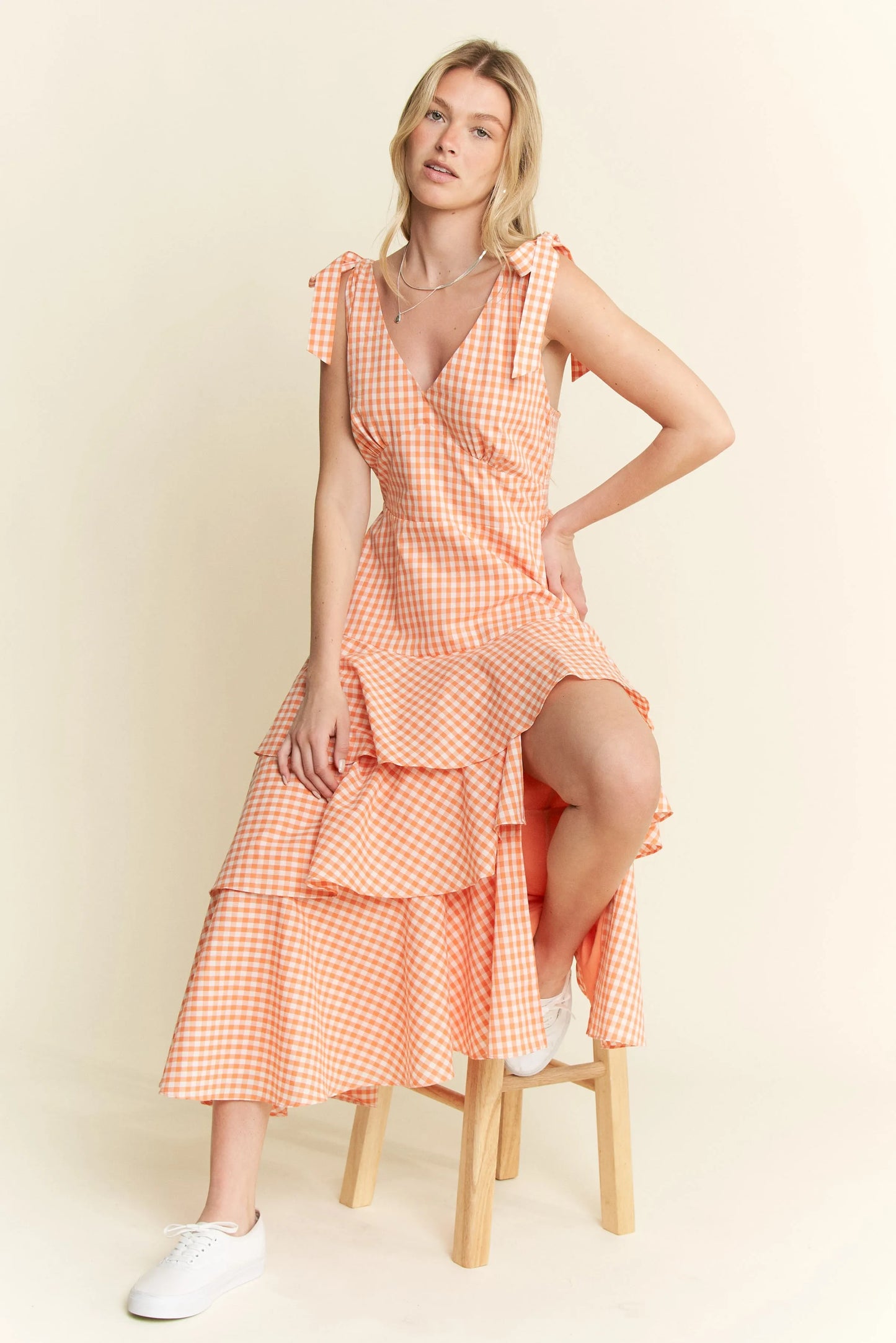 Seated front view of model wearing Alice Gingham Tiered Midi Dress by Jade by Jane, featuring V-neckline, tiered ruffle skirt, and orange gingham print.