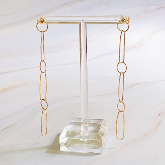Gold chain link earrings with textured detail hanging on clear stand