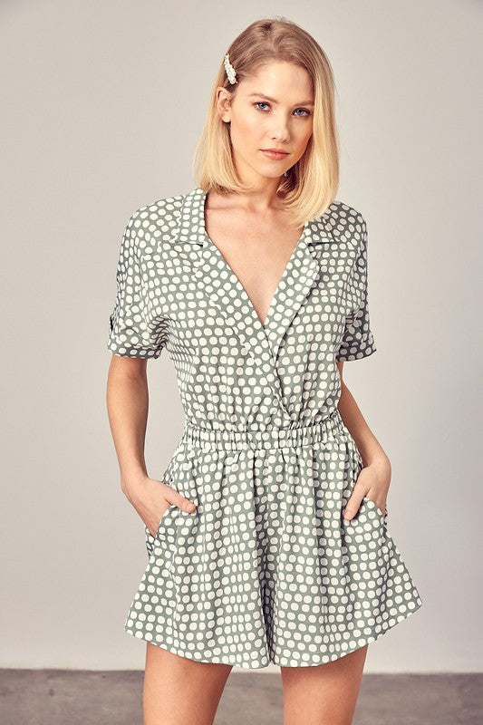 Sage-colored polka dot romper with a collared overlap neckline, short sleeves, and an elastic waist. Features a relaxed fit and a pocket.