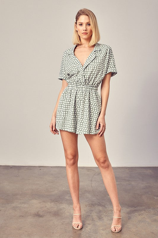 Sage-colored polka dot romper with a collared overlap neckline, short sleeves, and an elastic waist. Features a relaxed fit and a pocket.