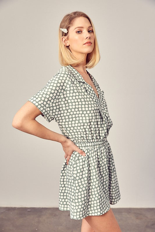 Sage-colored polka dot romper with a collared overlap neckline, short sleeves, and an elastic waist. Features a relaxed fit and a pocket.