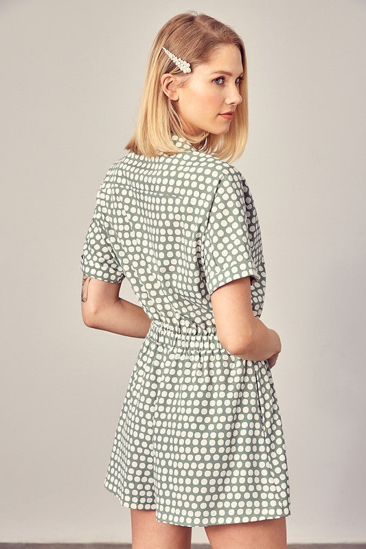 Sage-colored polka dot romper with a collared overlap neckline, short sleeves, and an elastic waist. Features a relaxed fit and a pocket.