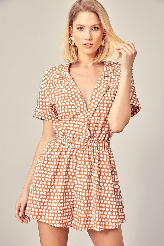 Clay-colored polka dot romper with a collared overlap neckline, short sleeves, and an elastic waist. Features a relaxed fit and a pocket.