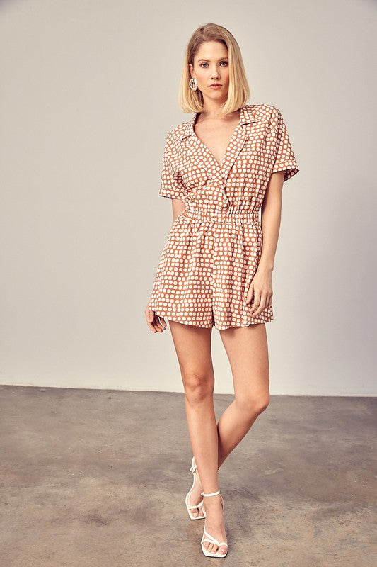 Clay-colored polka dot romper with a collared overlap neckline, short sleeves, and an elastic waist. Features a relaxed fit and a pocket.