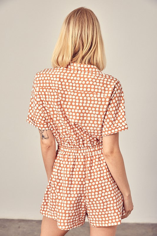 Clay-colored polka dot romper with a collared overlap neckline, short sleeves, and an elastic waist. Features a relaxed fit and a pocket.