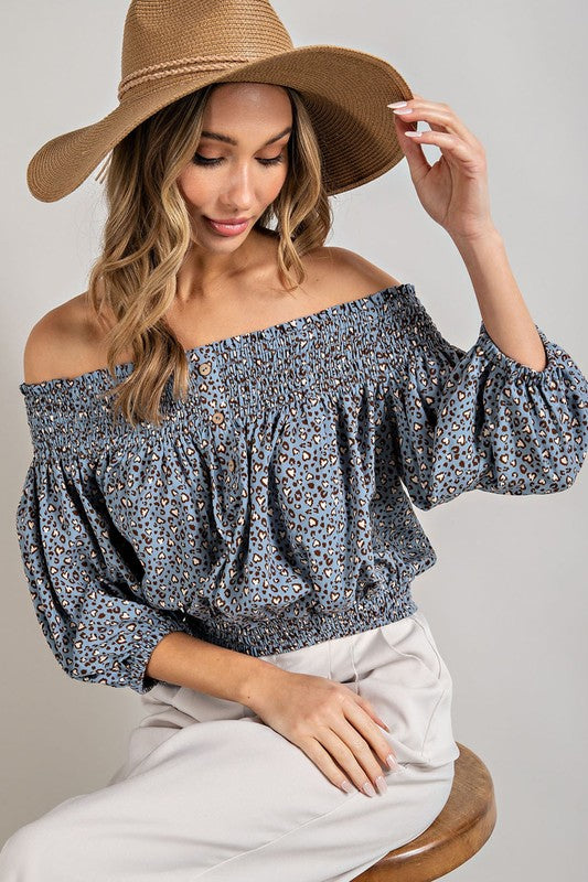 Blue animal print top with smocked off-the-shoulder neckline and button trim. Features a fitted and flared silhouette, 3/4 length puff sleeves, and a romantic, flirty style.