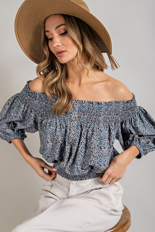 Blue animal print top with smocked off-the-shoulder neckline and button trim. Features a fitted and flared silhouette, 3/4 length puff sleeves, and a romantic, flirty style.