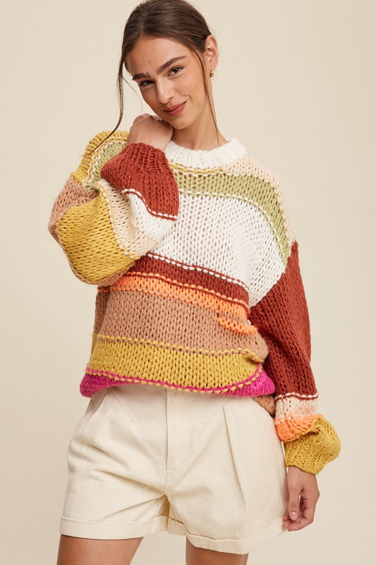 A cozy crochet sweater with a relaxed fit, featuring a round neckline accented with ribbed detailing. The long sleeves and hip-length silhouette create a comfortable and stylish look. The pullover design showcases vibrant stripes in shades of green, red, white, orange, and yellow, adding a playful and colorful touch.