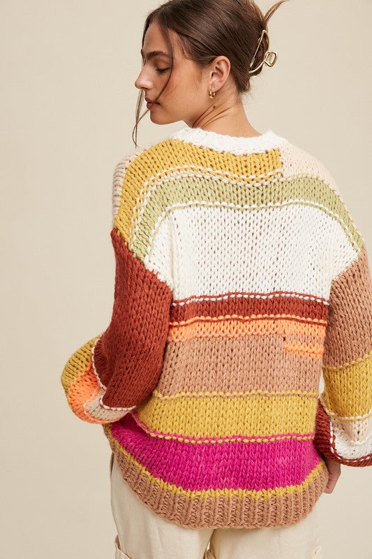 A cozy crochet sweater with a relaxed fit, featuring a round neckline accented with ribbed detailing. The long sleeves and hip-length silhouette create a comfortable and stylish look. The pullover design showcases vibrant stripes in shades of green, red, white, orange, and yellow, adding a playful and colorful touch.