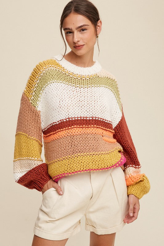 A cozy crochet sweater with a relaxed fit, featuring a round neckline accented with ribbed detailing. The long sleeves and hip-length silhouette create a comfortable and stylish look. The pullover design showcases vibrant stripes in shades of green, red, white, orange, and yellow, adding a playful and colorful touch.