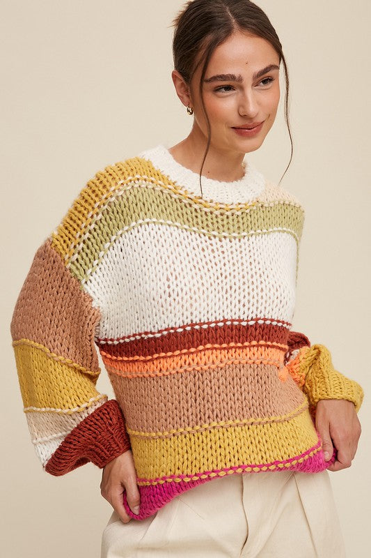 A cozy crochet sweater with a relaxed fit, featuring a round neckline accented with ribbed detailing. The long sleeves and hip-length silhouette create a comfortable and stylish look. The pullover design showcases vibrant stripes in shades of green, red, white, orange, and yellow, adding a playful and colorful touch.