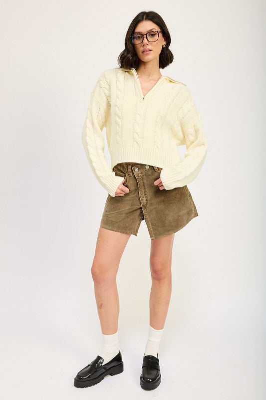 Cropped cable knit sweater in cream with a half-zip neckline, drop-shoulder long sleeves, and a ribbed hem.