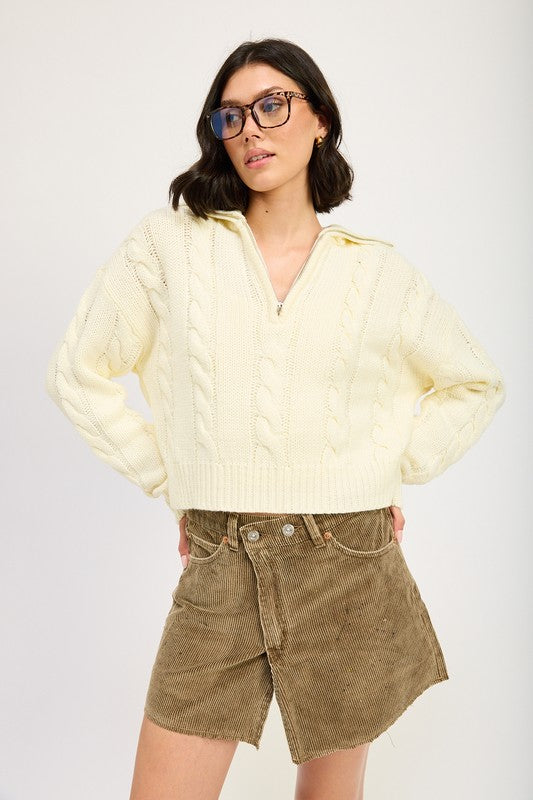 Cropped cable knit sweater in cream with a half-zip neckline, drop-shoulder long sleeves, and a ribbed hem.