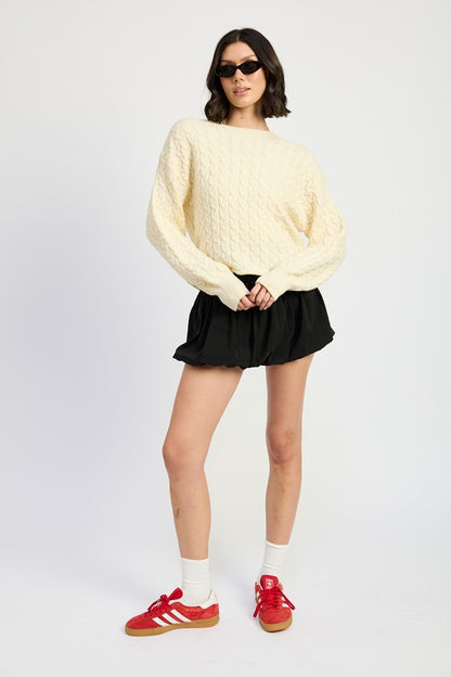 Full body view of model wearing ivory Kuuma Cable Knit Sweater with black skirt and red sneakers.