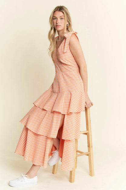 Side view of model seated in Alice Gingham Tiered Midi Dress by Jade by Jane, posing on chair with halter tie straps and flowy ruffle skirt, styled for a fresh summer look.