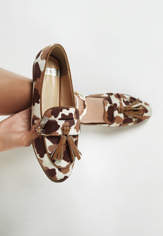 Model wearing one Moraug cowprint loafer and holding the other, showcasing faux suede texture, tassel detail, and stylish versatility by Rag Company