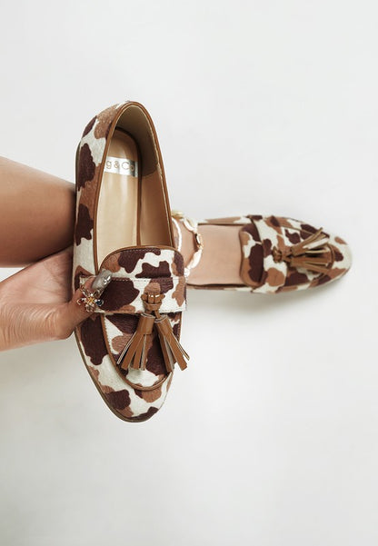 Model wearing one Moraug cowprint loafer and holding the other, showcasing faux suede texture, tassel detail, and stylish versatility by Rag Company