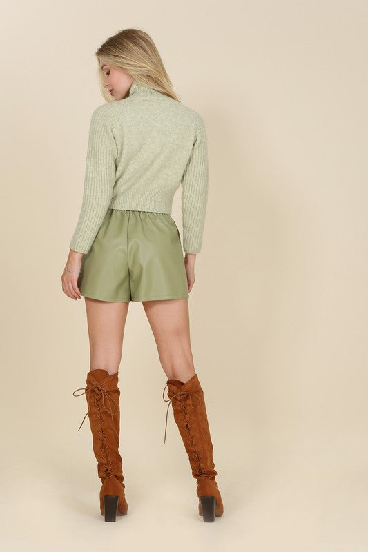 Trendy green vegan leather shorts featuring a hook and zipper closure, side pockets, pin-tuck detailing, and a comfortable wide elastic waistband.