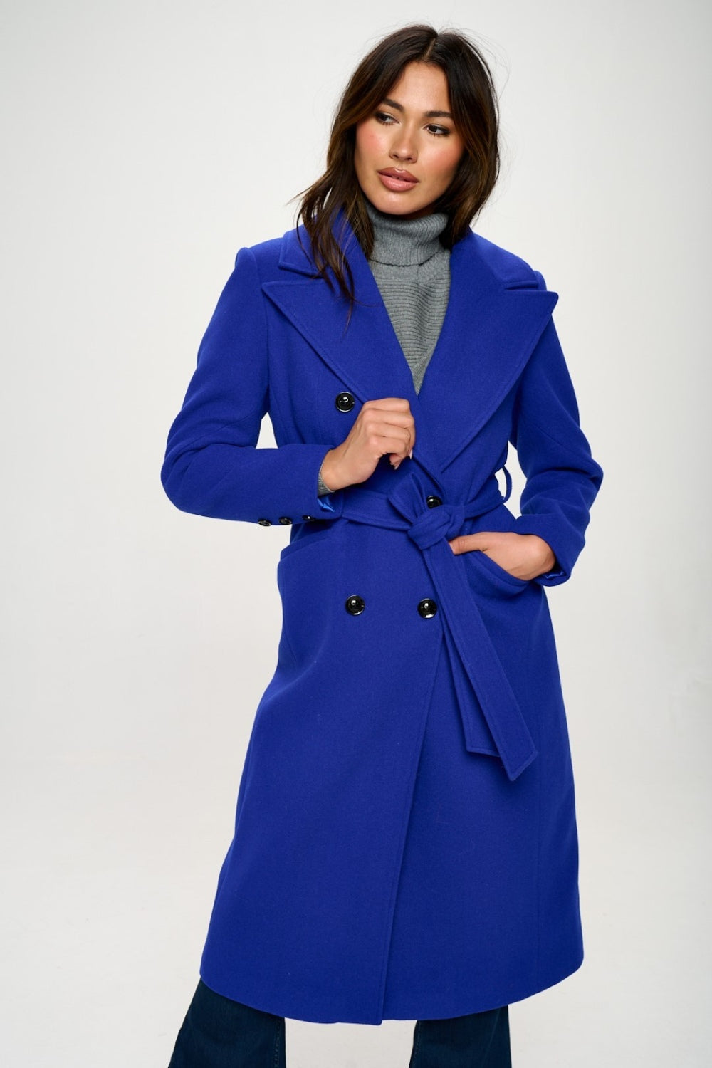 A royal blue double-breasted longline coat with a waist-cinching belt and long sleeves. The coat features a collared neckline, pocket details, and a tied closure.