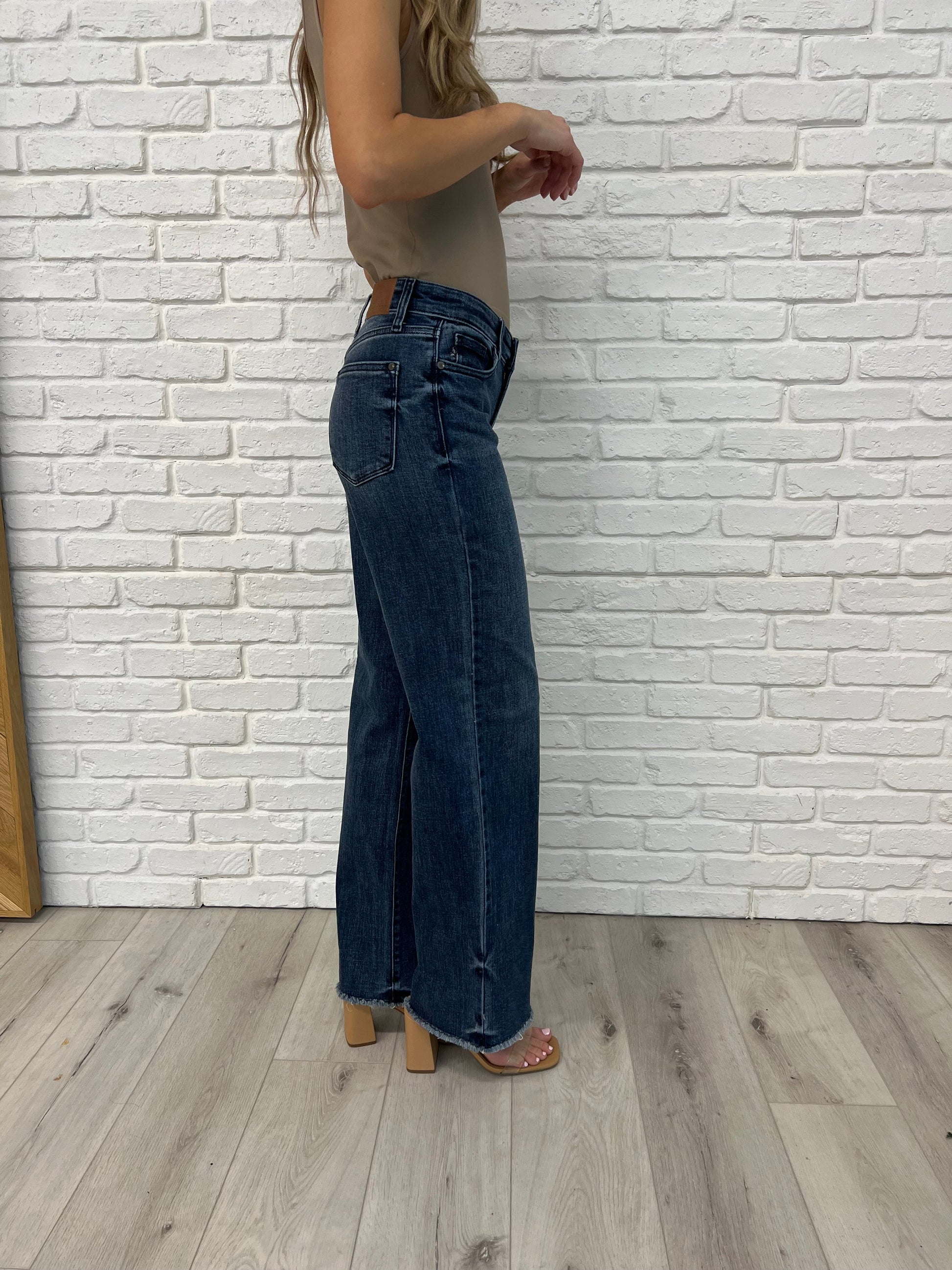 Side view of the Addie Mid Rise Wide Leg Jeans by Judy Blue in dark vintage wash, featuring raw cut-off hem, release hem dye detail, and a 93% cotton blend for a retro-inspired look with modern flair.