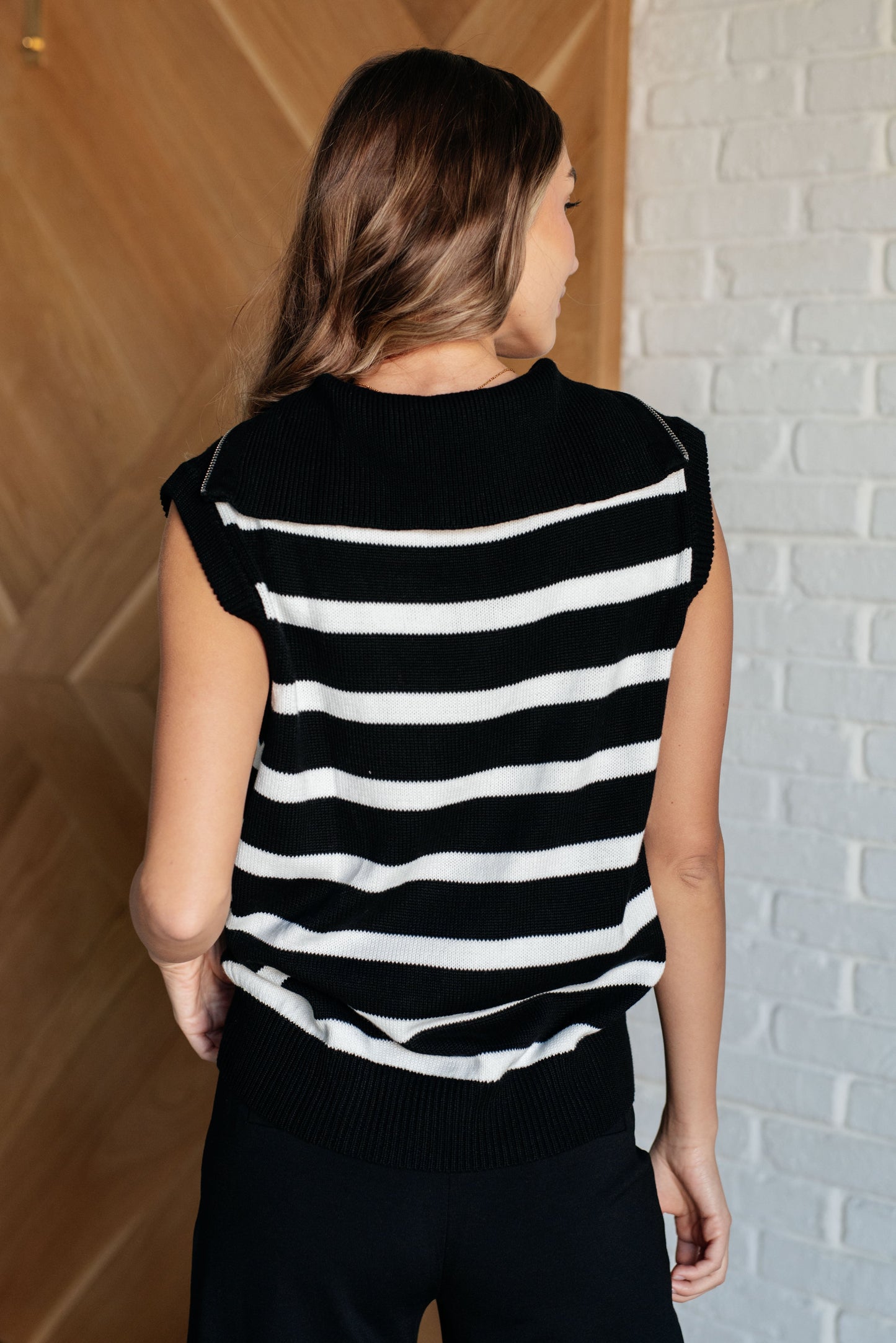 Black and white sleeveless sweater knit top featuring a striped pattern, a collared neckline, and a half-zip closure. It includes banded ribbed cuffs and a hemline for added detail.