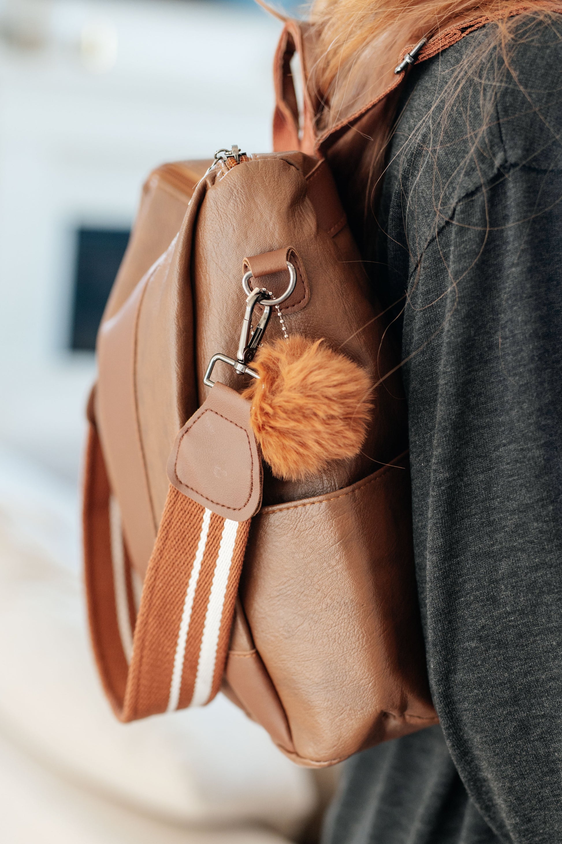 Brown vegan leather women's backpack with a fluffy ball key charm, removable canvas strap, fixed handle, backpack straps, and a zippered closure. Features include side pockets, a back side zip pocket, and an inside pocket with dark silver hardware accents.