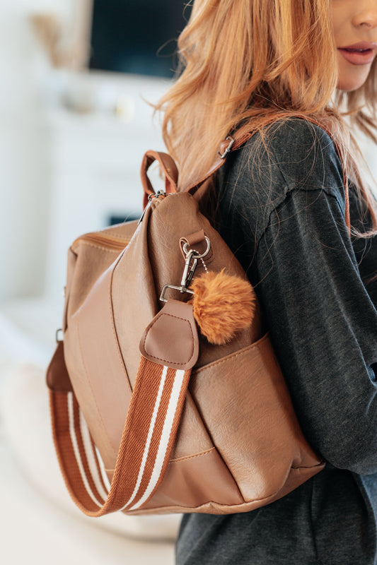 Brown vegan leather women's backpack with a fluffy ball key charm, removable canvas strap, fixed handle, backpack straps, and a zippered closure. Features include side pockets, a back side zip pocket, and an inside pocket with dark silver hardware accents.