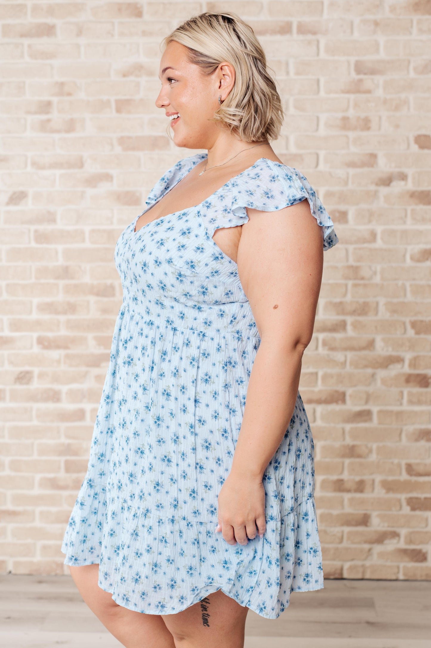 McKay V-Neck Floral Dress in blue with a playful floral print, featuring a flattering V-neckline, flutter sleeves, open back with a tie detail, and crinkle woven fabric for effortless charm. Fully lined for comfort, perfect for brunches, picnics, or special occasions.