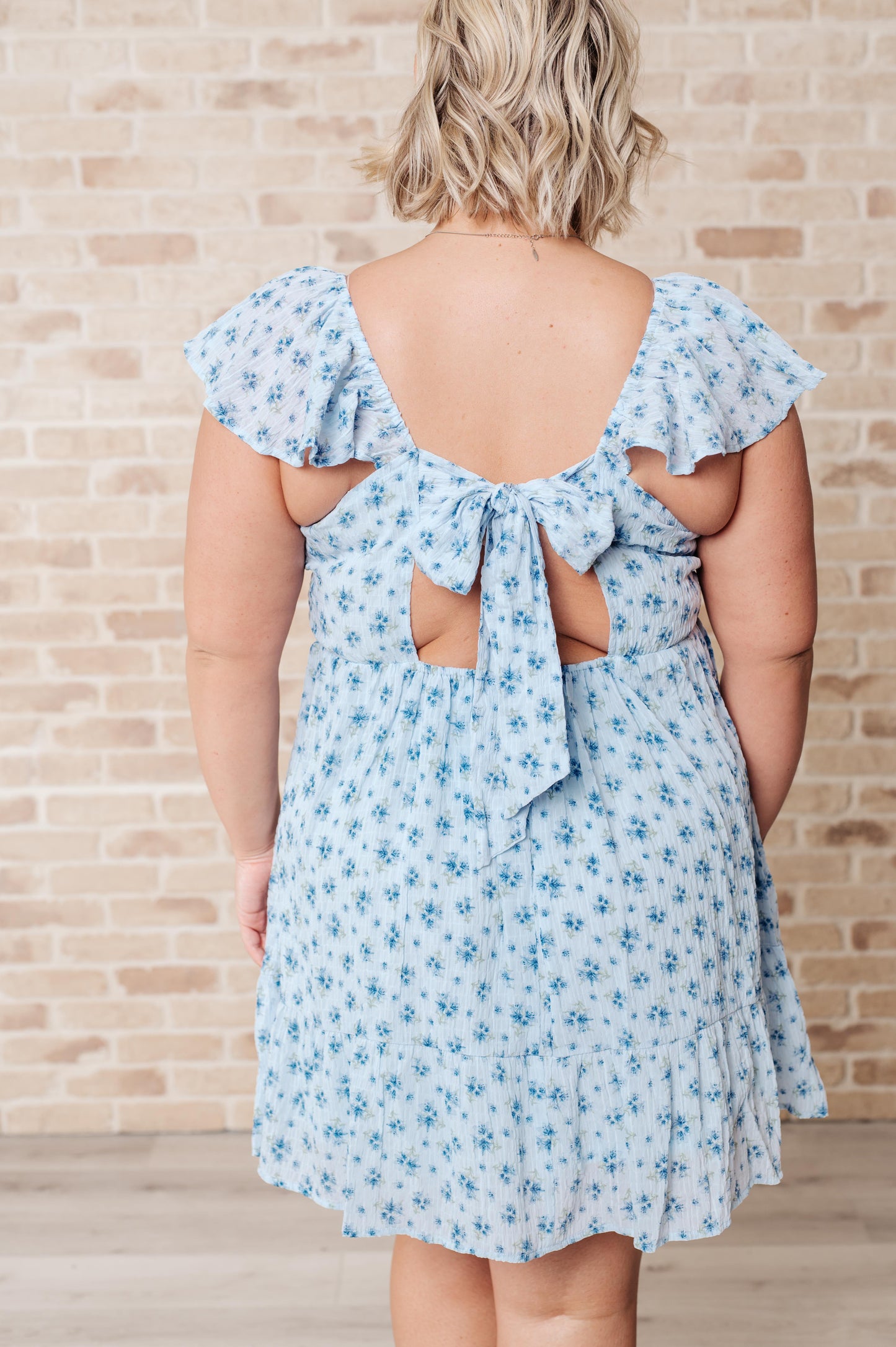 McKay V-Neck Floral Dress in blue with a playful floral print, featuring a flattering V-neckline, flutter sleeves, open back with a tie detail, and crinkle woven fabric for effortless charm. Fully lined for comfort, perfect for brunches, picnics, or special occasions.