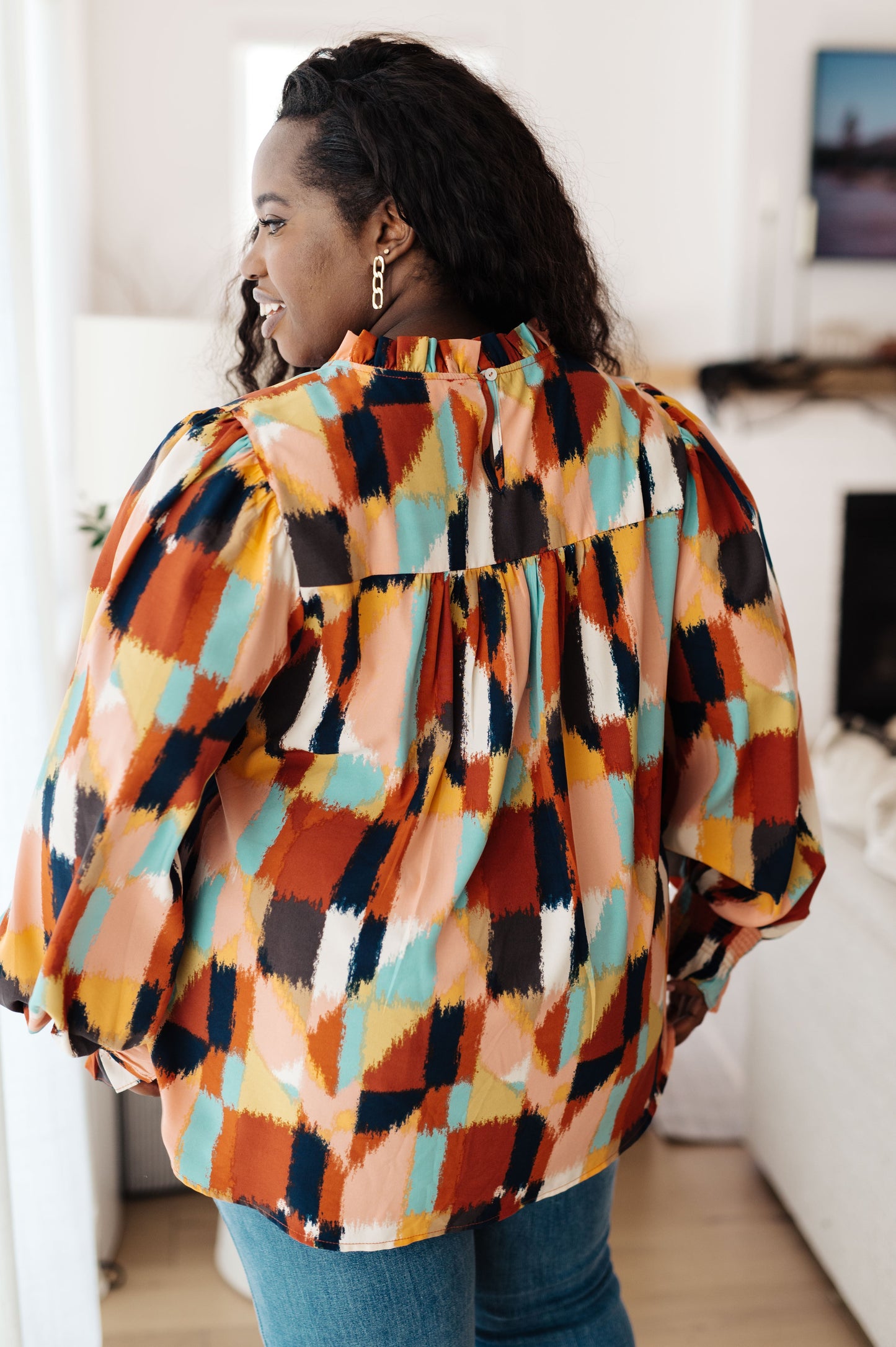 Back view of the plus size model wearing the Padme Blouse featuring a bold mod print, ruffled neckline, and smocked elastic sleeve cuffs for a chic, vintage-inspired look with a modern twist.