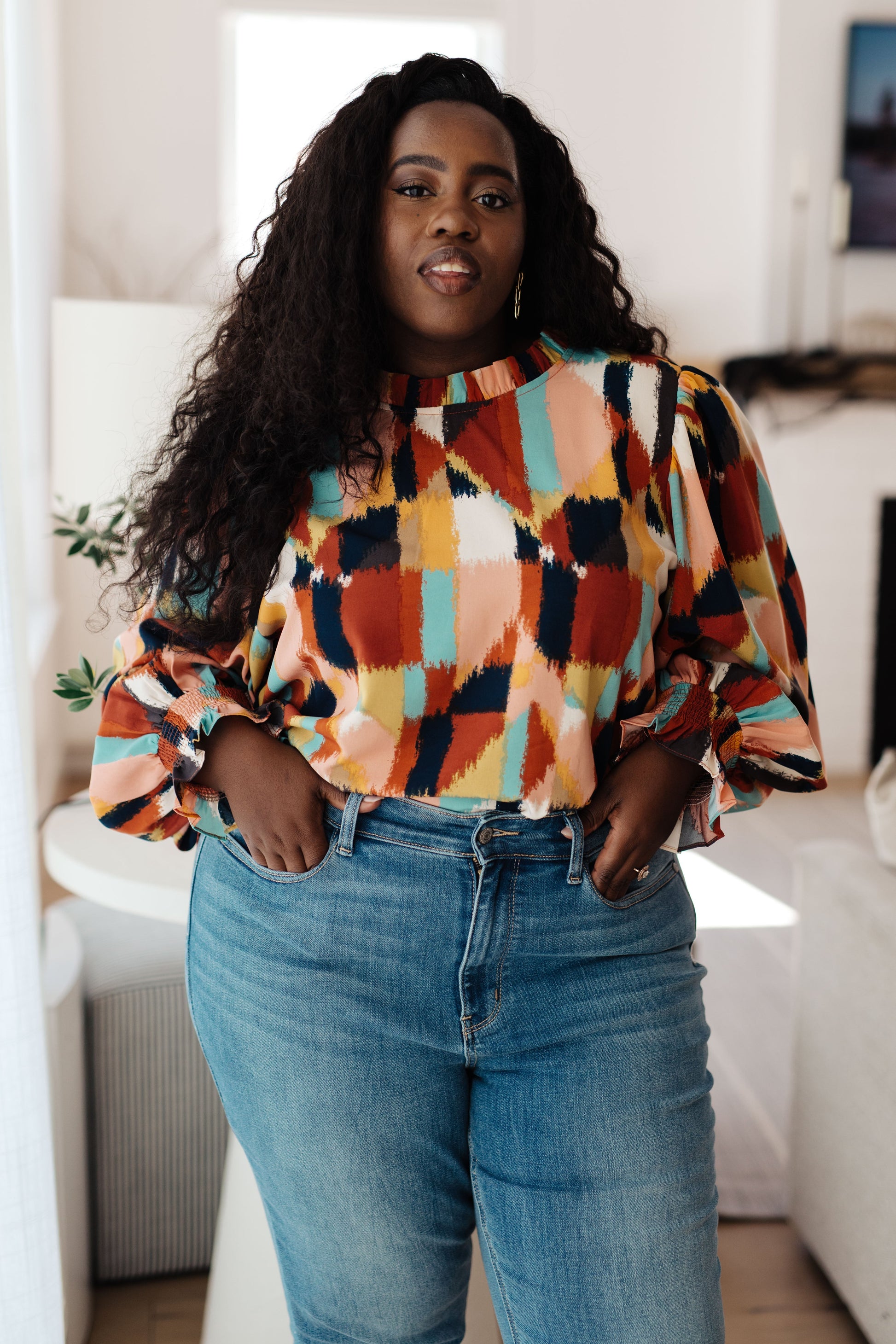 Plus size model wearing the Padme Blouse featuring a bold mod print, ruffled neckline, and smocked elastic sleeve cuffs for a chic, vintage-inspired look with a modern twist.