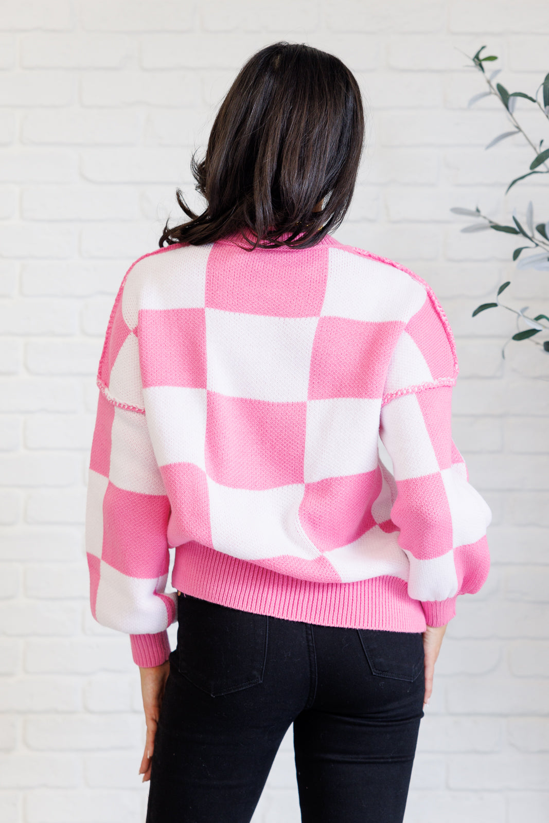 A women's pink and white checkered sweater with a round neckline, long sleeves with drop shoulders, exaggerated ribbed cuffs, exposed seam details, and a banded ribbed hemline.