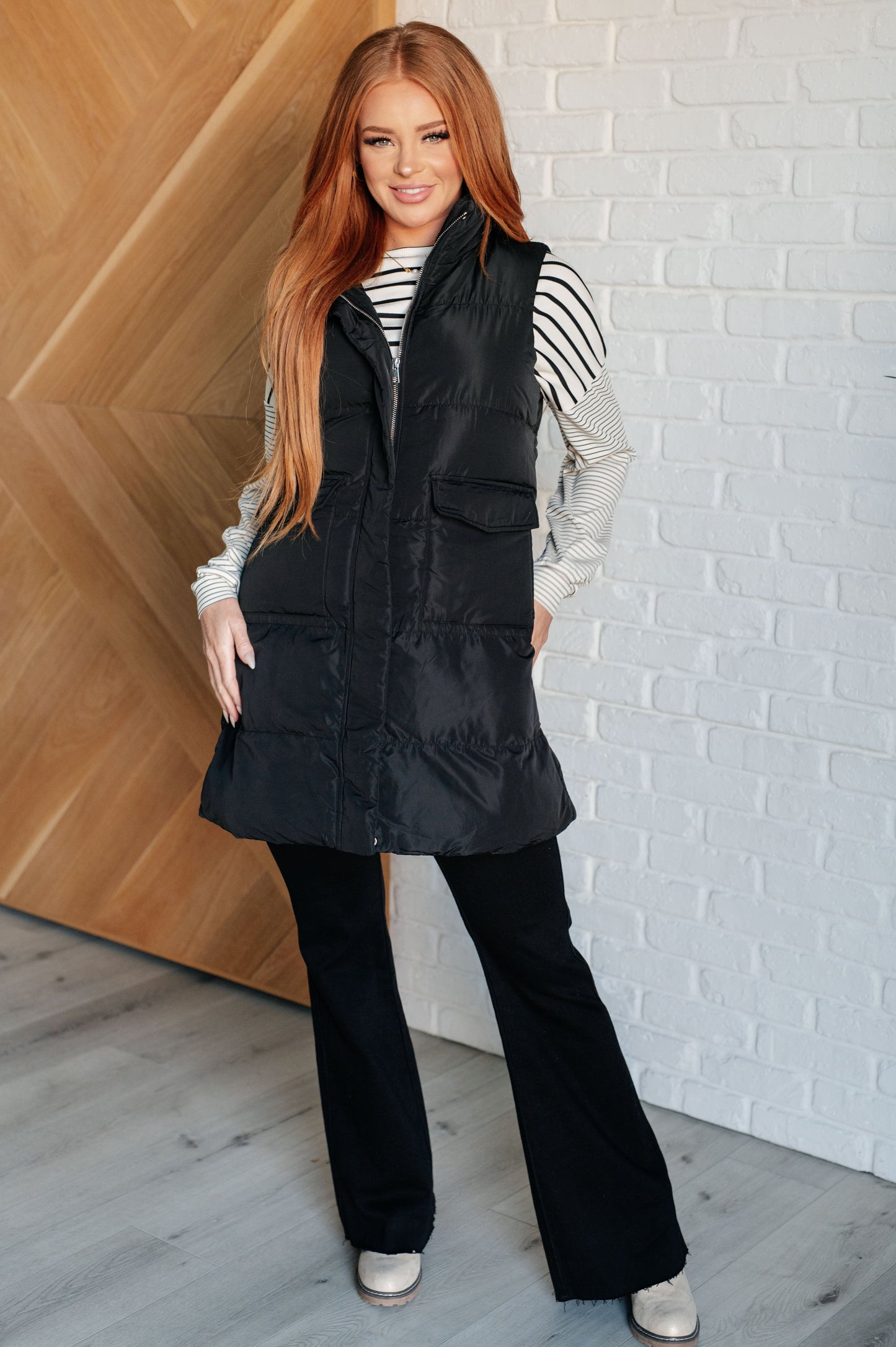 A women's longline puffer vest in black, featuring a high neckline, sleeveless design, full front zipper closure with buttons, and patch pockets. Made from woven windbreaker material.