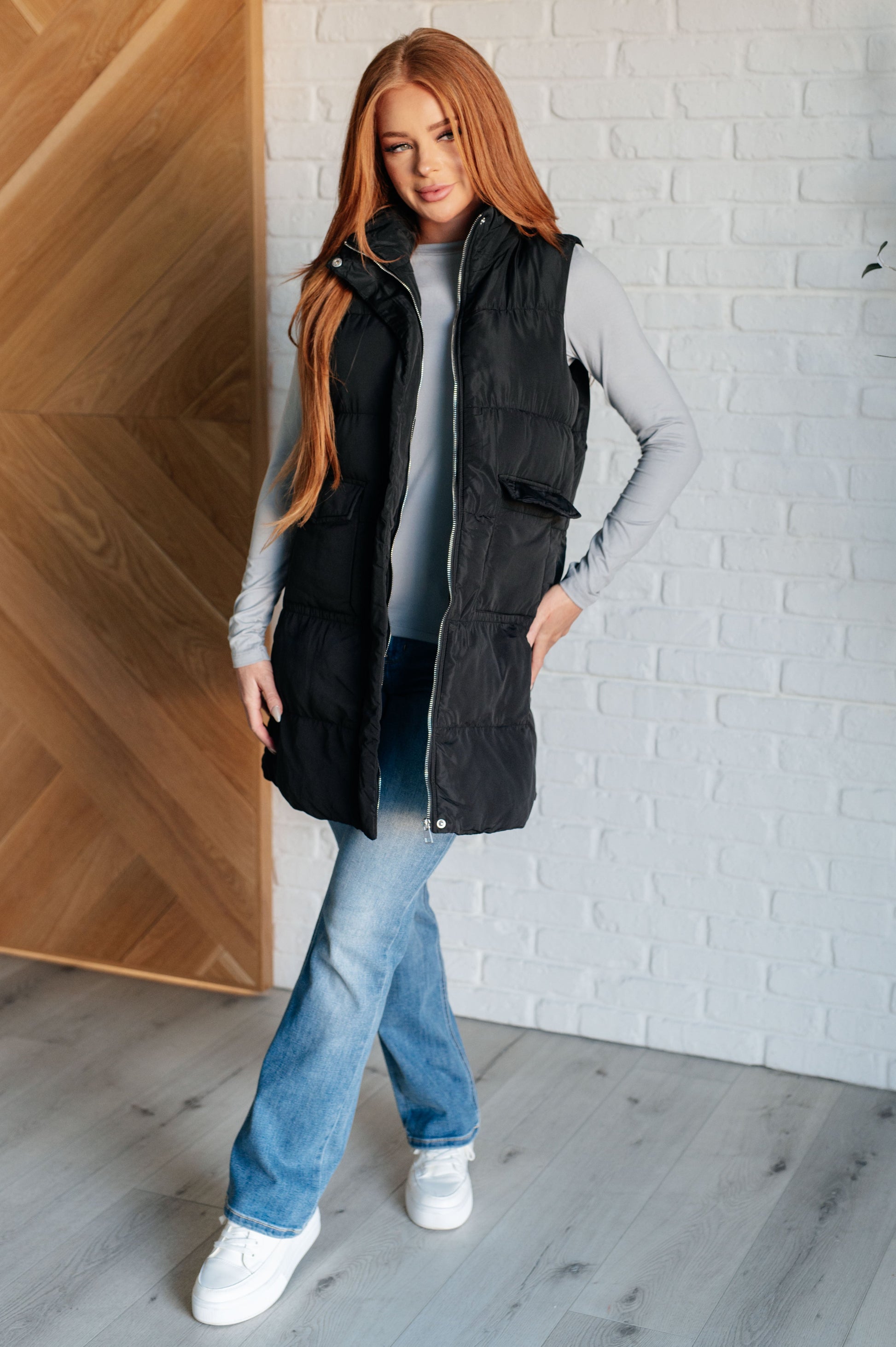 A women's longline puffer vest in black, featuring a high neckline, sleeveless design, full front zipper closure with buttons, and patch pockets. Made from woven windbreaker material.