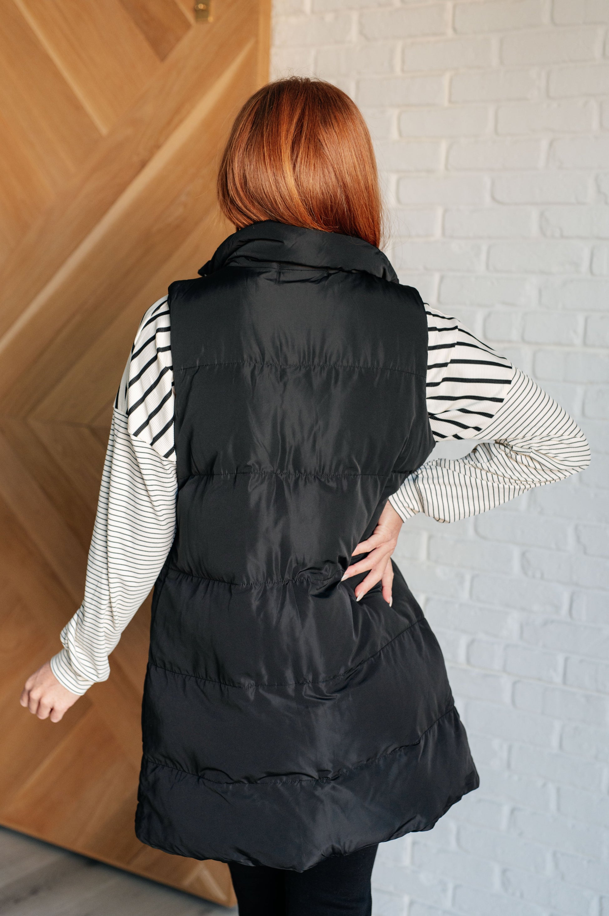 A women's longline puffer vest in black, featuring a high neckline, sleeveless design, full front zipper closure with buttons, and patch pockets. Made from woven windbreaker material.