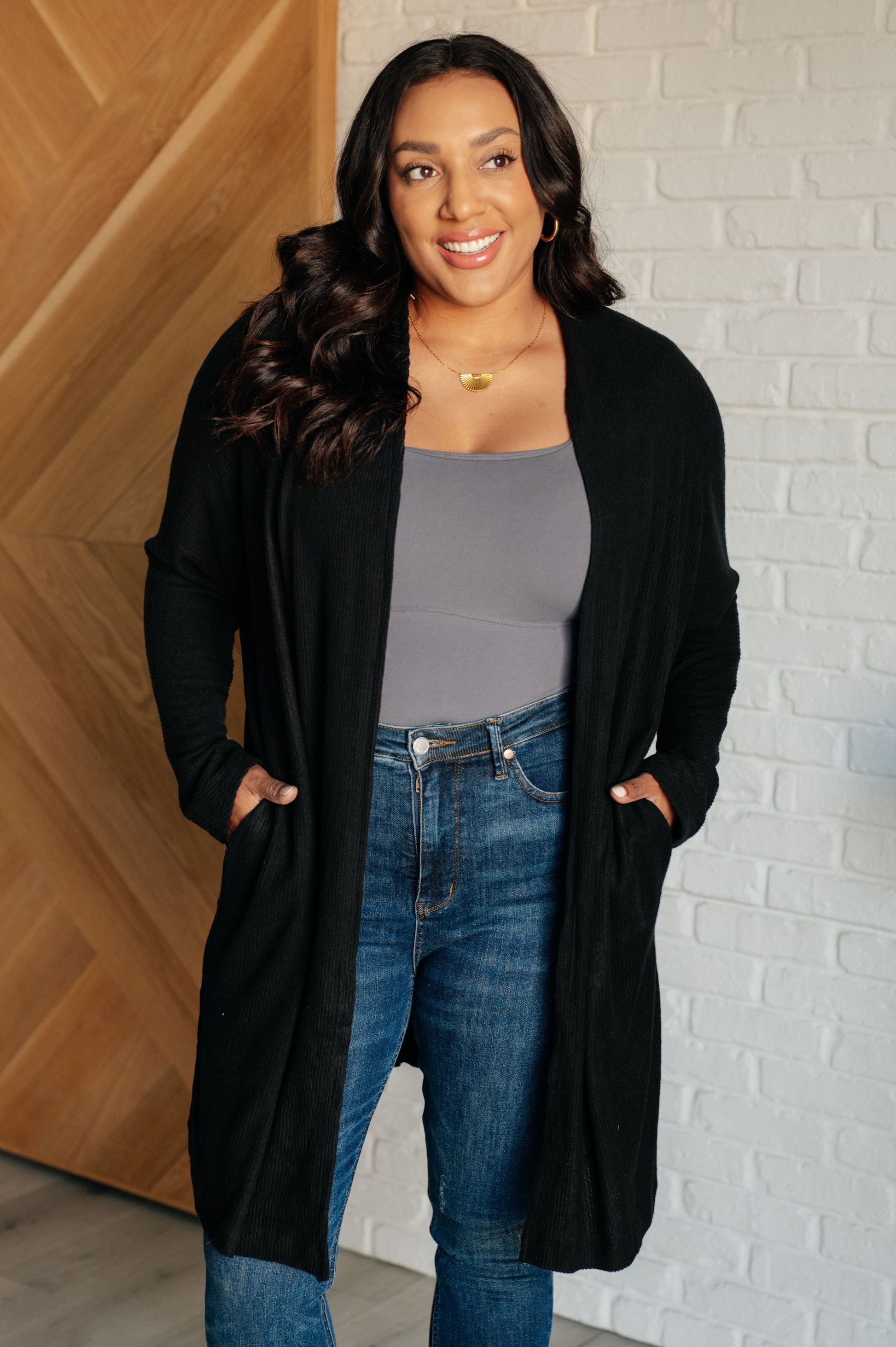 Black ribbed knit cardigan with a shawl neckline and long dolman sleeves. The open-front design features a long length, functional side pockets, and subtle side slits.