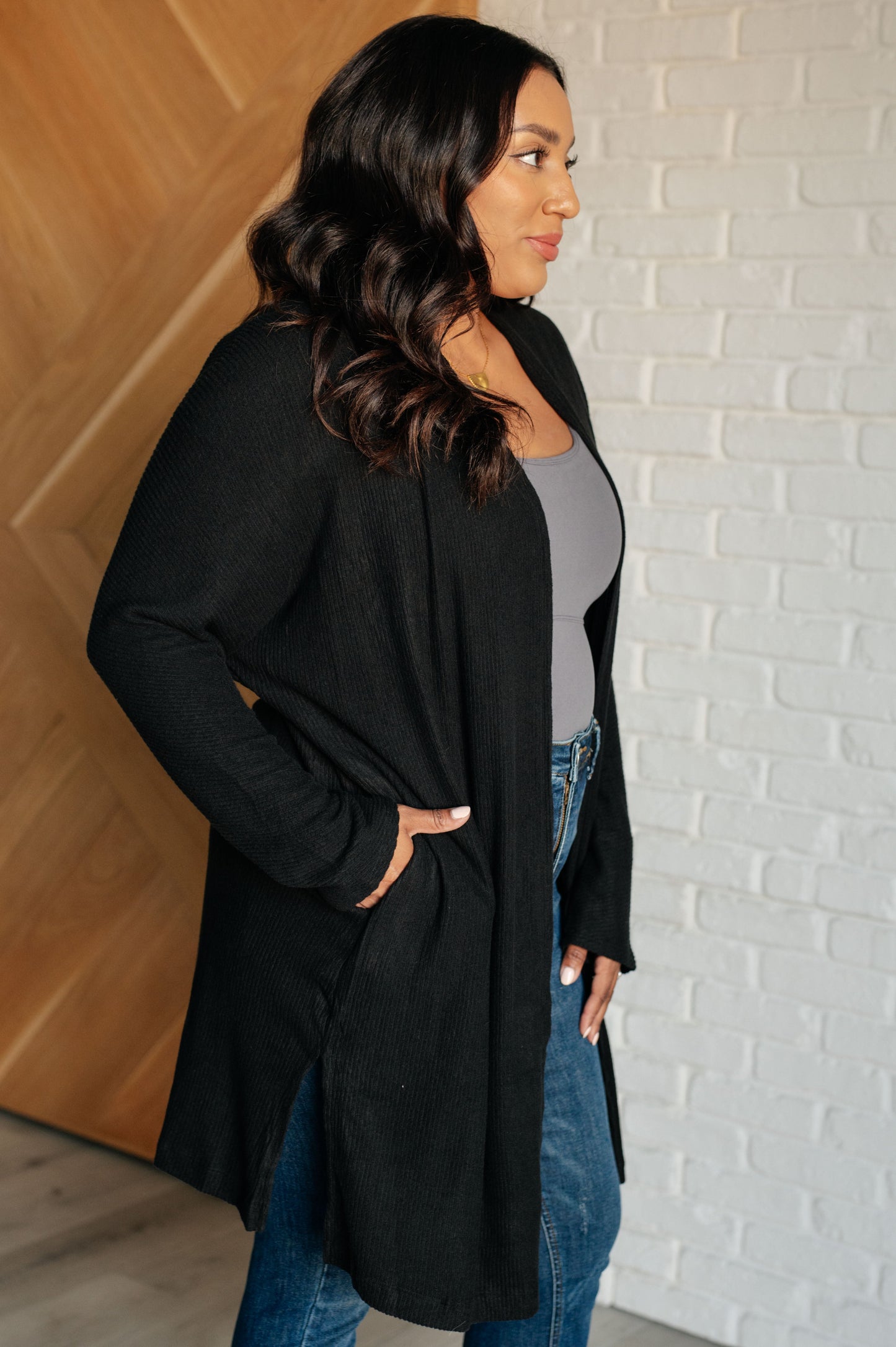 Black ribbed knit cardigan with a shawl neckline and long dolman sleeves. The open-front design features a long length, functional side pockets, and subtle side slits.