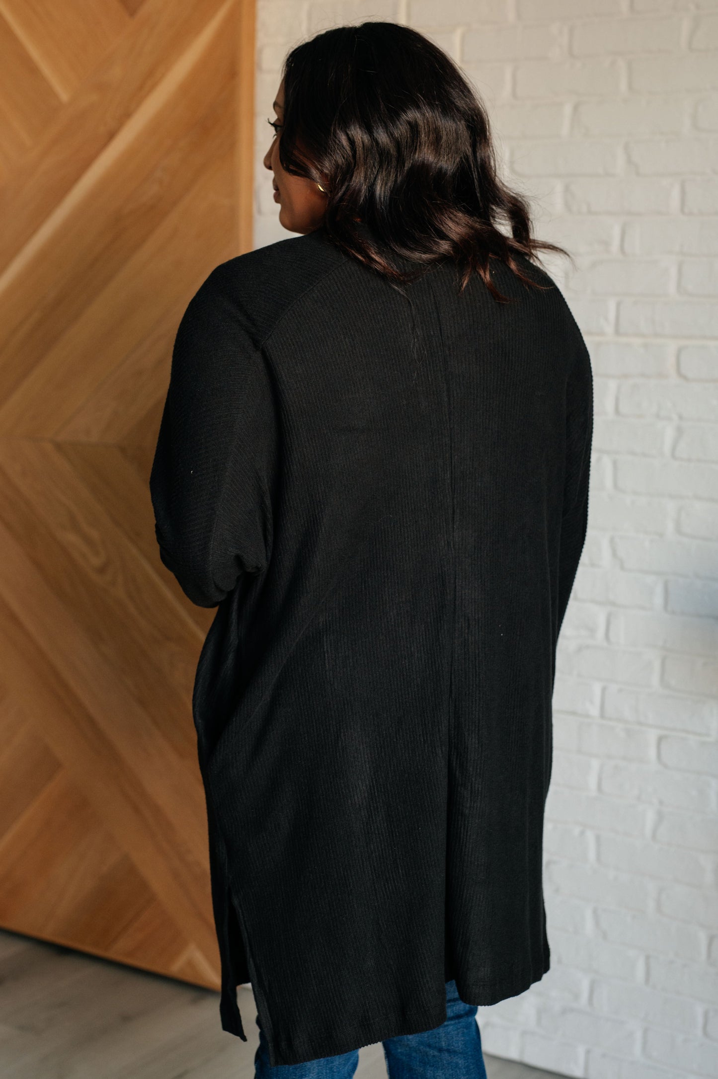 Black ribbed knit cardigan with a shawl neckline and long dolman sleeves. The open-front design features a long length, functional side pockets, and subtle side slits.