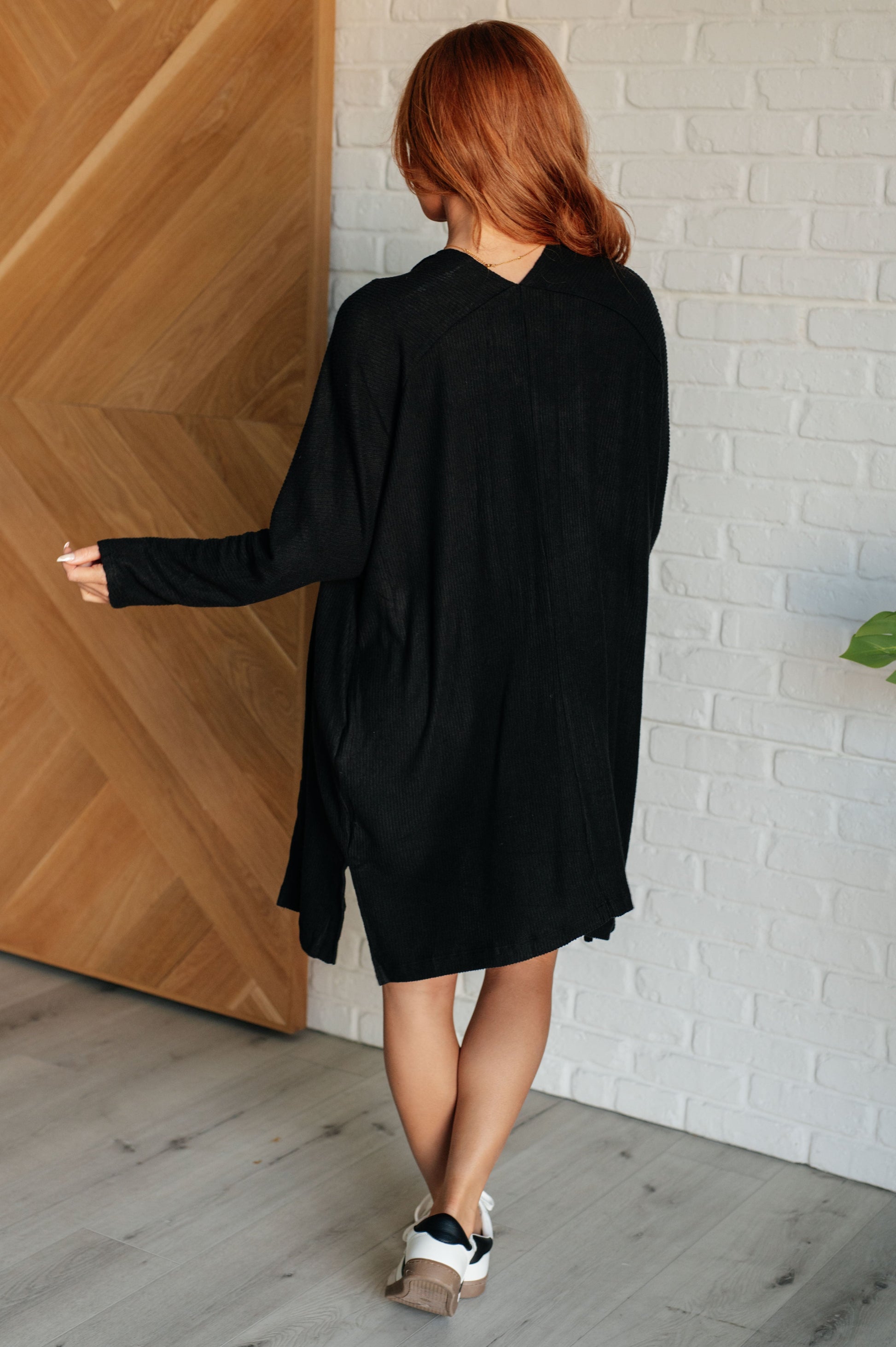 Black ribbed knit cardigan with a shawl neckline and long dolman sleeves. The open-front design features a long length, functional side pockets, and subtle side slits.