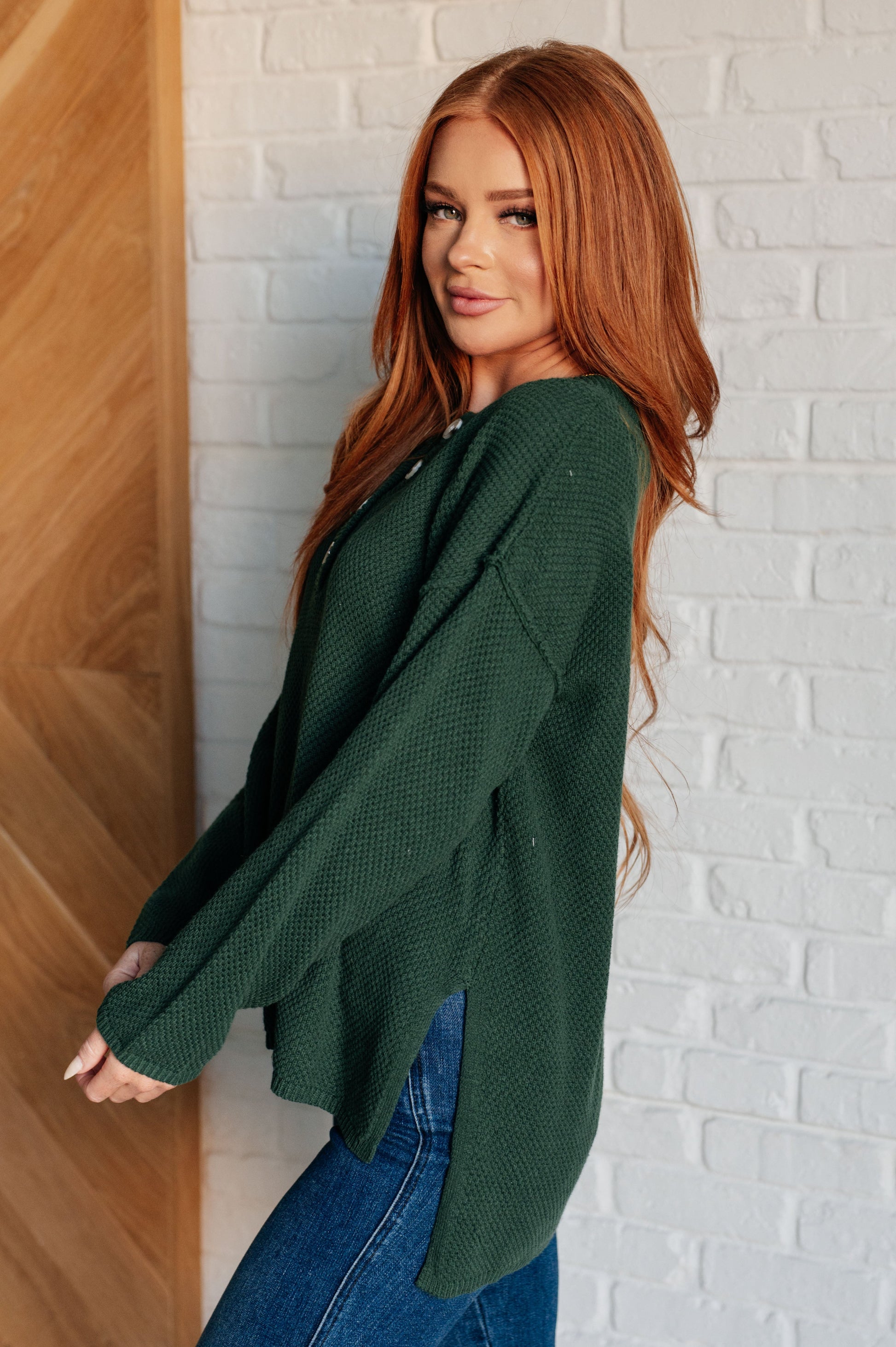 Women's dark green henley sweater with a ribbed button-up neckline, exposed seams, long sleeves, and a stepped hem with side slits.