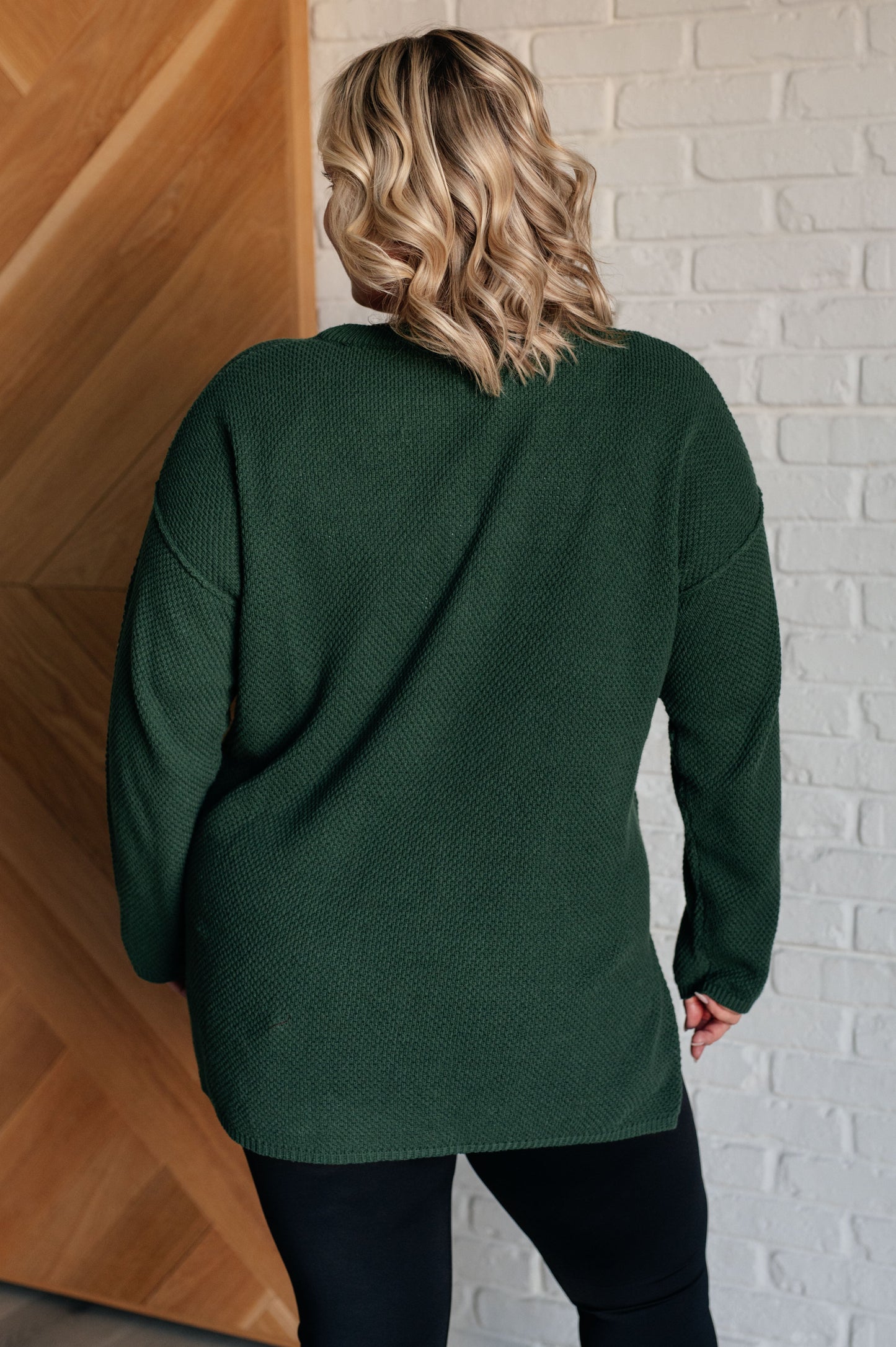 Women's dark green henley sweater with a ribbed button-up neckline, exposed seams, long sleeves, and a stepped hem with side slits.